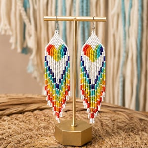 May include: A pair of beaded earrings with a rainbow heart design. The earrings are white with a colorful pattern of red, orange, yellow, green, blue, and purple beads. The earrings are displayed on a gold-colored stand.