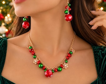 May include: A gold-tone chain necklace and matching drop earrings featuring red and green jingle bells, small red and green spheres with white snowflake patterns, and gold-tone snowflake charms. The jewelry set is worn against a dark green top.