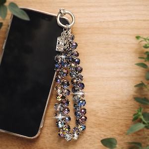 May include: A silver-toned phone charm with a beaded design. The charm features iridescent, multi-colored beads in shades of blue, purple, and gold. The charm is attached to a silver ring and a small rectangular tag. The charm is next to a smartphone.