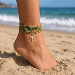 May include: Anklet with green and gold beads, featuring a gold palm leaf charm. The beaded anklet is worn on a foot on a sandy beach, with the ocean in the background. The anklet is a summer accessory.