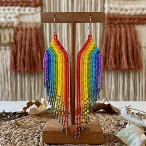 May include: Rainbow beaded earrings with a vibrant spectrum of colors, including red, orange, yellow, green, blue, and purple. The earrings are displayed on a wooden stand, showcasing their intricate design and beaded fringe.