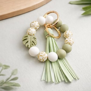 May include: A beaded keychain bracelet in shades of white, green, and gold. The bracelet features a gold keyring, a light green tassel, and a decorative leaf. The beads have a leopard print design.