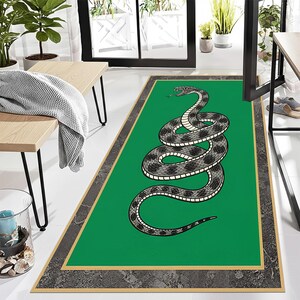Puede incluir: Una alfombra rectangular verde con un diseño de serpiente enroscada en tonos grises y negros. La alfombra tiene un borde gris oscuro con un ribete dorado. La lengua de la serpiente está extendida. La alfombra está sobre un suelo blanco.