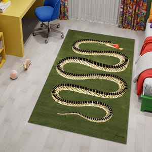 May include: Green rectangular rug with a stylized snake design. The snake has a black and white pattern and a red tongue. The rug is on a light wood floor in a room with a desk and chair.