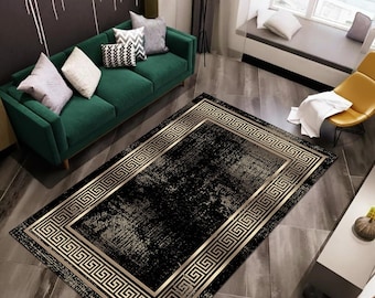 May include: A rectangular black area rug with a gold Greek key border. The rug has a distressed, textured appearance. The rug is placed on a dark gray floor in a living room setting. The rug is a decorative home decor item.