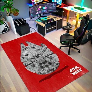 May include: A red rectangular rug with a detailed grayscale image of the Millennium Falcon starship from Star Wars. The Star Wars logo is in the bottom right corner. The rug is on a light wood floor.