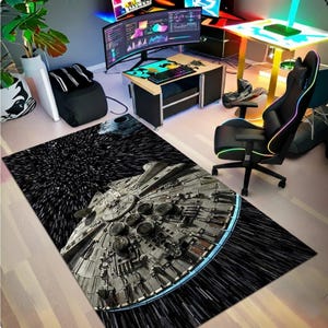 Puede incluir: Una alfombra rectangular con una imagen detallada de la nave espacial Millennium Falcon en un campo estelar. La alfombra tiene un fondo negro con rayas blancas, y la nave espacial es gris con acentos azules. La alfombra está colocada sobre un suelo de madera clara.