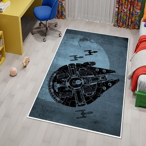 May include: A rectangular rug featuring a black silhouette of the Millennium Falcon spaceship against a blue background with a large circular shape and three smaller spacecraft. The rug has a white border and is placed on a light wood floor.