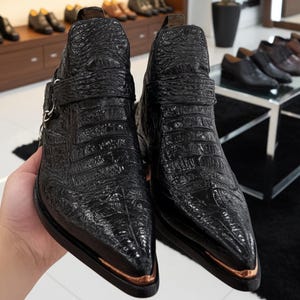 Könnte beinhalten: Ein Paar schwarze Leder-Ankle-Boots mit Krokodiltextur. Die Stiefel haben spitze Zehen mit kupferfarbenen Metallakzenten und einen Riemen mit einer silbernen Schnalle. Weitere Schuhe sind im Hintergrund zu sehen.