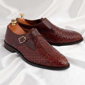 May include: A pair of dark brown leather monk strap shoes featuring a woven texture on the vamp and side panels. Each shoe has a smooth leather heel counter, a gold-toned metal buckle closure, and a wooden shoe tree inserted inside the opening.