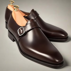 Könnte beinhalten: Ein Paar polierte braune Lederschuhe mit einem einzelnen Riemen und einer silbernen Schnalle. Die Schuhe haben ein klassisches Design mit glänzender Oberfläche. Schuhspanner aus Holz sind eingesetzt. Ca. 30 cm lang.