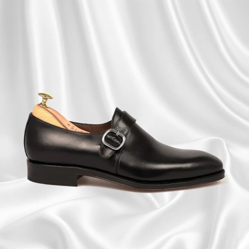 May include: A black leather single monk strap shoe featuring a silver-toned metal buckle closure across the instep. The shoe includes a light-colored wooden shoe tree inserted inside the heel area, resting on a smooth, solid black sole.