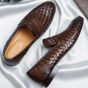 Op de afbeelding: Een paar bruine leren loafers met een geweven design. De schoenen hebben een klassieke instapstijl met een band over de bovenkant. De zolen zijn donkerbruin en de binnenkant is lichtbruin.