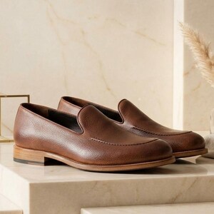 May include: A pair of brown leather loafers with a textured finish. The shoes have a classic slip-on design with a rounded toe and a low wooden heel. The loafers are displayed on a light-colored surface.