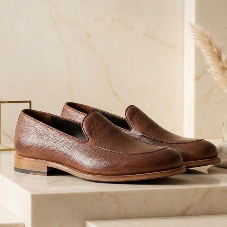 May include: A pair of brown leather loafers with a textured finish. The shoes have a classic slip-on design with a rounded toe and a low wooden heel. The loafers are displayed on a light-colored surface.