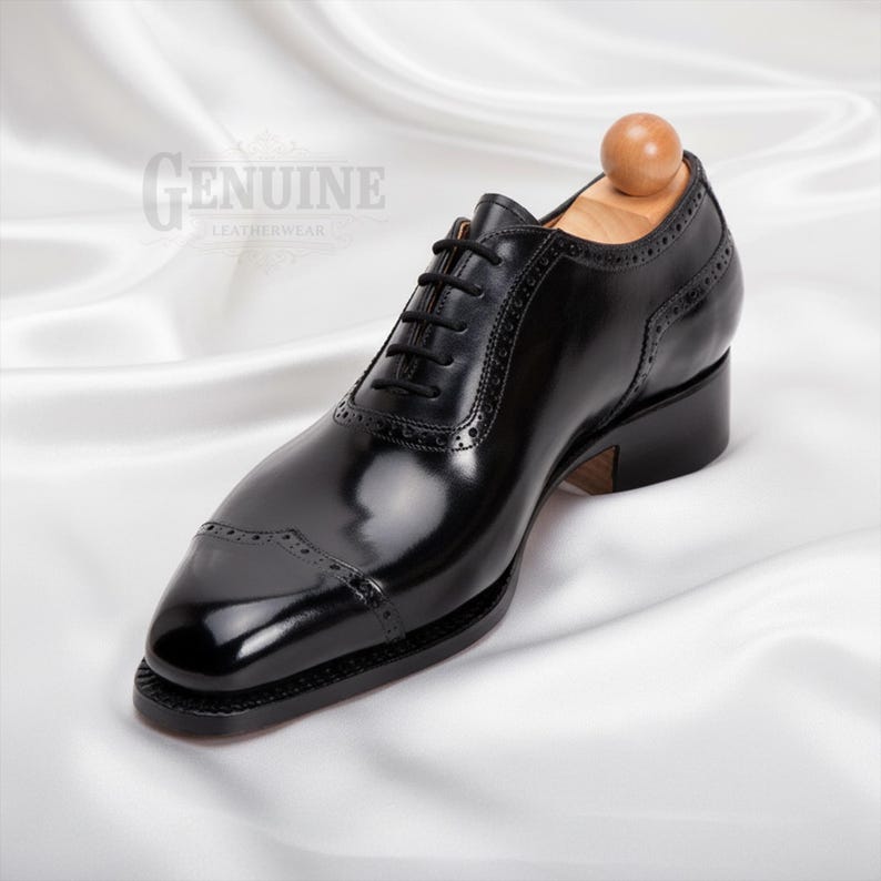 Puede incluir: Un zapato oxford de cuero negro pulido con puntera decorada con perforaciones brogue, cordones negros y una horma de madera. El lateral del zapato muestra el texto Genuine Leatherwear en un estilo de fuente decorativo.