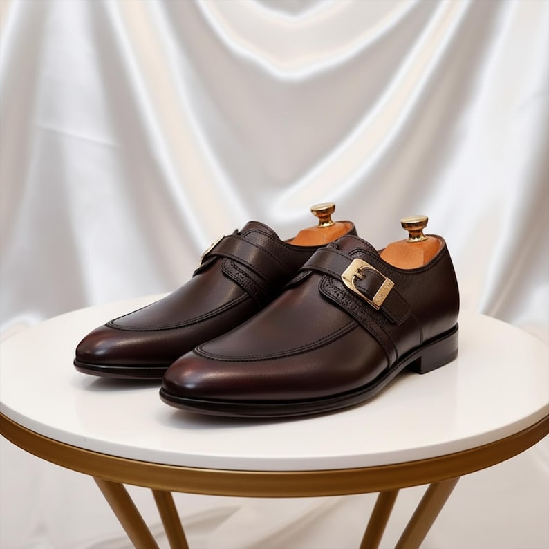 Pode incluir: Um par de sapatos de couro castanho com fecho de fivela. Os sapatos t&ecirc;m um design elegante com bico fino e fivela dourada. Os sapatos s&atilde;o exibidos numa mesa branca com base dourada.