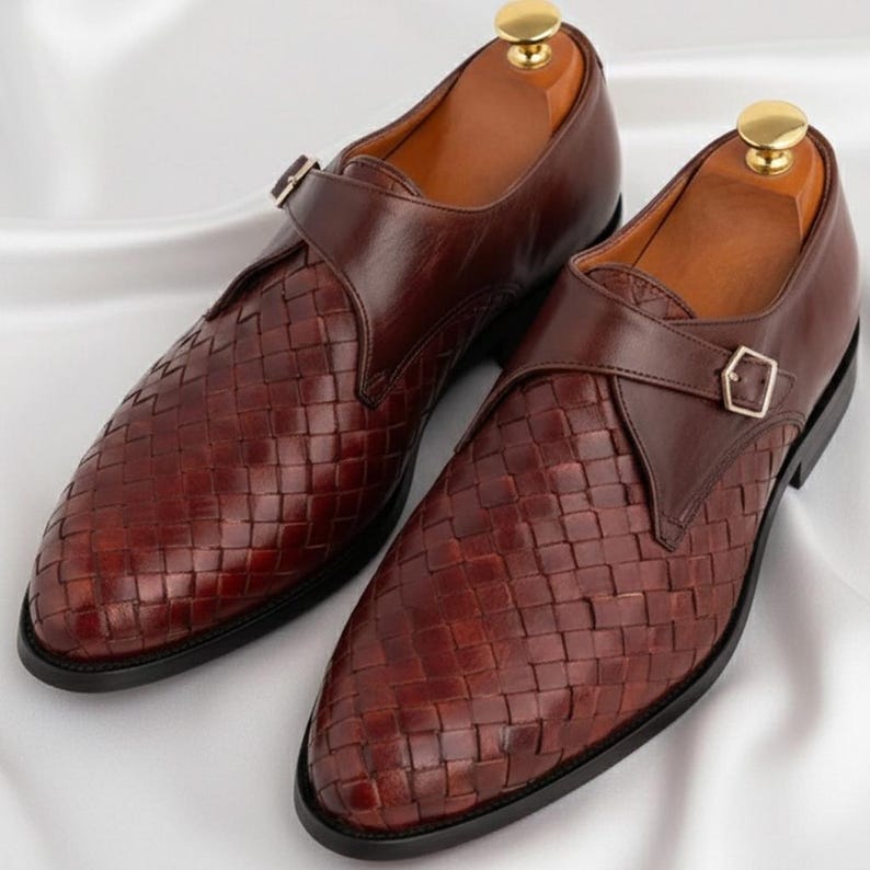 May include: A pair of dark brown leather monk strap shoes featuring a woven texture on the vamp and toe box. Each shoe has a smooth leather strap with a gold-toned metal buckle, a black sole, and a wooden shoe tree inserted inside the opening.