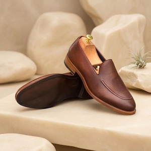 May include: A pair of brown leather loafers with a textured finish. The shoes have a classic slip-on design with a rounded toe and a wooden shoe tree inside. The soles are a darker brown, and the shoes are set against a neutral background.
