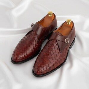 May include: A pair of dark brown leather monk strap shoes featuring a woven texture on the vamp and a single gold-toned buckle closure. The shoes have a smooth leather finish on the heel and strap area, with a gold-colored shoe tree insert.