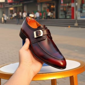 Pode incluir: Um sapato de couro bord&ocirc; com fivela de tira &uacute;nica. O sapato apresenta perfura&ccedil;&otilde;es decorativas nas bordas e bico fino. O sapato &eacute; segurado por uma m&atilde;o, mostrando o acabamento polido e o design.