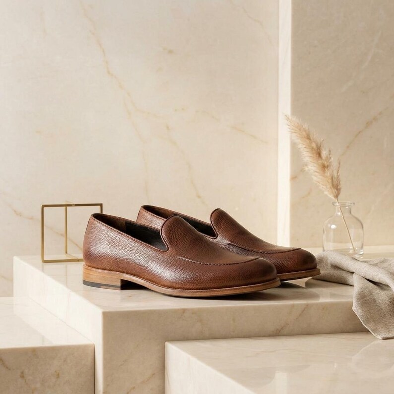 May include: A pair of brown leather loafers with a textured finish. The shoes have a classic slip-on design with a low heel and are displayed on a marble surface. The background features a neutral color palette.