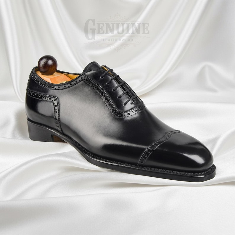Puede incluir: Un zapato oxford de cuero negro pulido con puntera reforzada y detalles decorativos de brogue, cordones negros finos y una horma de madera. El zapato tiene grabado el texto GENUINE LEATHERWEAR cerca de la zona del tal&oacute;n.