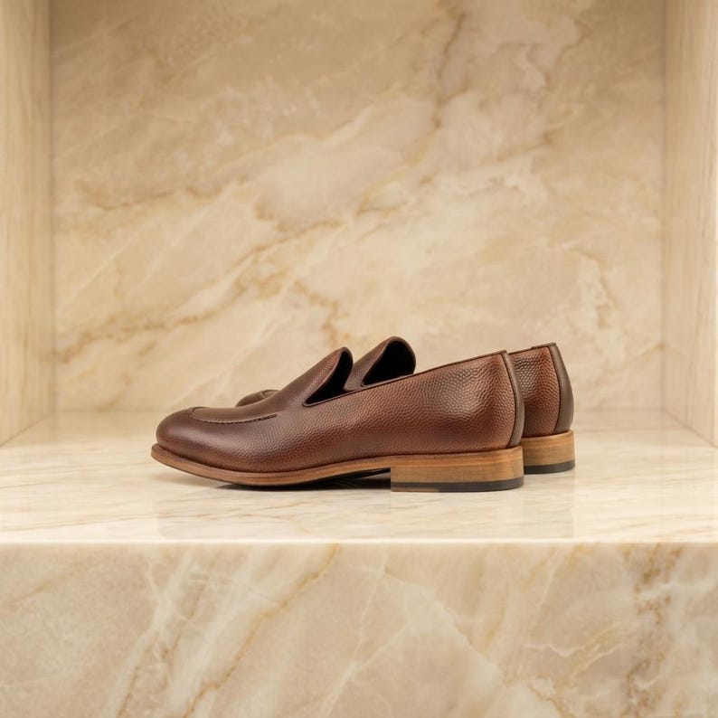 May include: A pair of brown leather loafers with a textured finish. The shoes have a classic slip-on design with a low heel and a light brown sole. The shoes are displayed on a marble shelf.