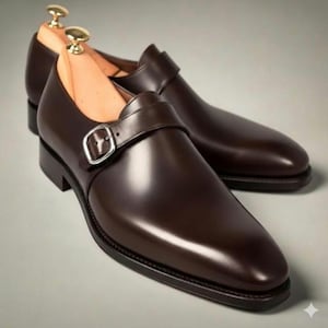 May include: A pair of dark brown leather single monk strap shoes with a polished finish, featuring a silver-toned metal buckle closure and a wooden shoe tree inserted into each shoe to maintain the structured shape of the footwear.