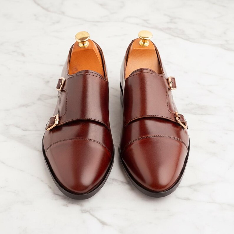 K&ouml;nnte beinhalten: Ein Paar polierte, dunkelbraune Doppel-Monk-Schuhe aus Leder mit Zehenkappe, zwei goldfarbenen Metallschnallen an jedem Schuh und hellen Holzschuhspannern, die im Inneren eingesetzt sind, um die Form des Schuhwerks beizubehalten.