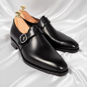 May include: A pair of black leather single monk strap shoes with a polished finish, featuring a silver-toned metal buckle closure and a rounded toe. Each shoe contains a light-colored wooden shoe tree inserted inside to maintain its shape.