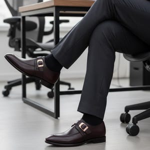 Pode incluir: Sapatos sociais de couro castanho com fivelas douradas, usados com cal&ccedil;as cinza escuro e meias pretas. Sapatos de estilo cl&aacute;ssico, adequados para ocasi&otilde;es formais.
