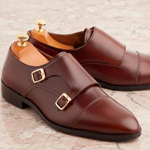 K&ouml;nnte beinhalten: Ein Paar dunkelbraune Lederschuhe mit Doppel-Monk-Riemen, Zehenkappe, goldfarbenen Metallschnallen und in jeden Schuh eingesetzten Schuhspannern aus Holz. Die Schuhe haben eine glatte Oberfl&auml;che und eine dunkelbraune Sohle mit einem kleinen Absatz.