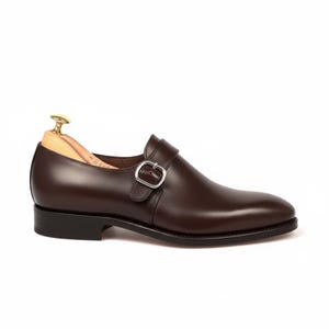 May include: A dark brown leather single monk strap shoe featuring a silver-toned metal buckle, a smooth rounded toe, and a low black heel. A light-colored wooden shoe tree is inserted inside the shoe to maintain its shape and structure.