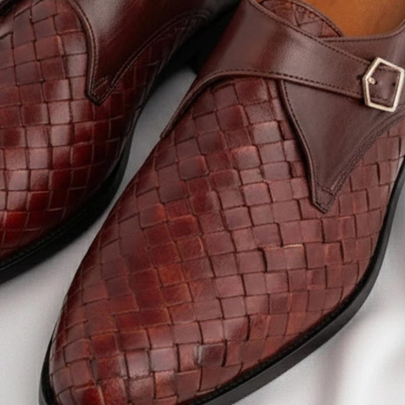 May include: A pair of dark brown leather monk strap shoes featuring a woven basketweave pattern across the vamp and toe. Each shoe includes a smooth leather strap secured with a polished silver-toned metal buckle over the instep area.