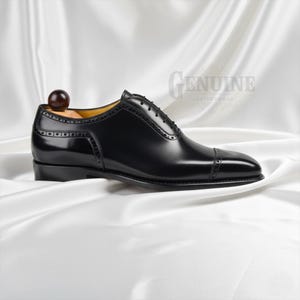 Puede incluir: Un zapato oxford de cuero negro con puntera reforzada y detalles decorativos de brogue a lo largo de las costuras y los paneles de los ojales. El zapato incluye una horma de madera con un pomo redondo en el tal&oacute;n y cordones negros atados.