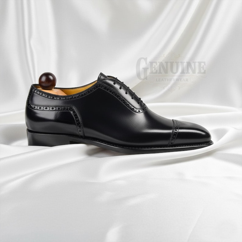 Puede incluir: Un zapato oxford de cuero negro con puntera reforzada y detalles decorativos de brogue a lo largo de las costuras y los paneles de los ojales. El zapato incluye una horma de madera con un pomo redondo en el tal&oacute;n y cordones negros atados.