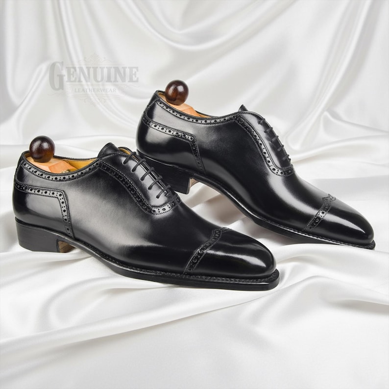 Puede incluir: Un par de zapatos oxford de cuero negro con puntera y detalles decorativos de brogue, cordones negros finos y hormas de madera insertadas en el interior. La palabra GENUINE est&aacute; impresa en la esquina superior izquierda de la imagen.