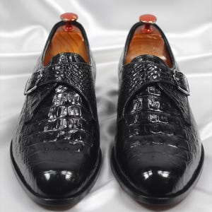 May include: A pair of black shoes with a glossy, textured finish resembling crocodile skin. Each shoe features a single monk strap with a silver-toned metal buckle across the top. The interior of the shoes shows a light brown wooden shoe tree.