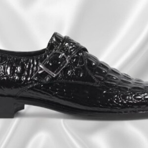 May include: A black shoe with a glossy finish and a textured crocodile skin pattern. The shoe features a single monk strap across the top with a silver-toned metal buckle and a low, solid black heel. The design includes a rounded toe profile.