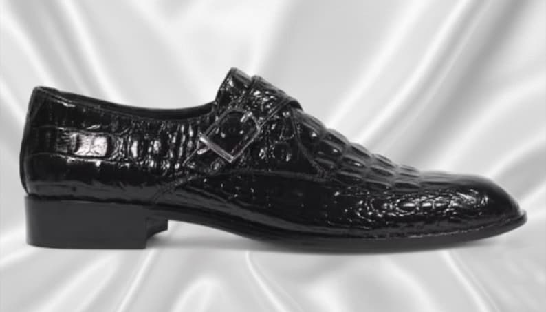 May include: A black shoe with a glossy finish and a textured crocodile skin pattern. The shoe features a single monk strap across the top with a silver-toned metal buckle and a low, solid black heel. The design includes a rounded toe profile.