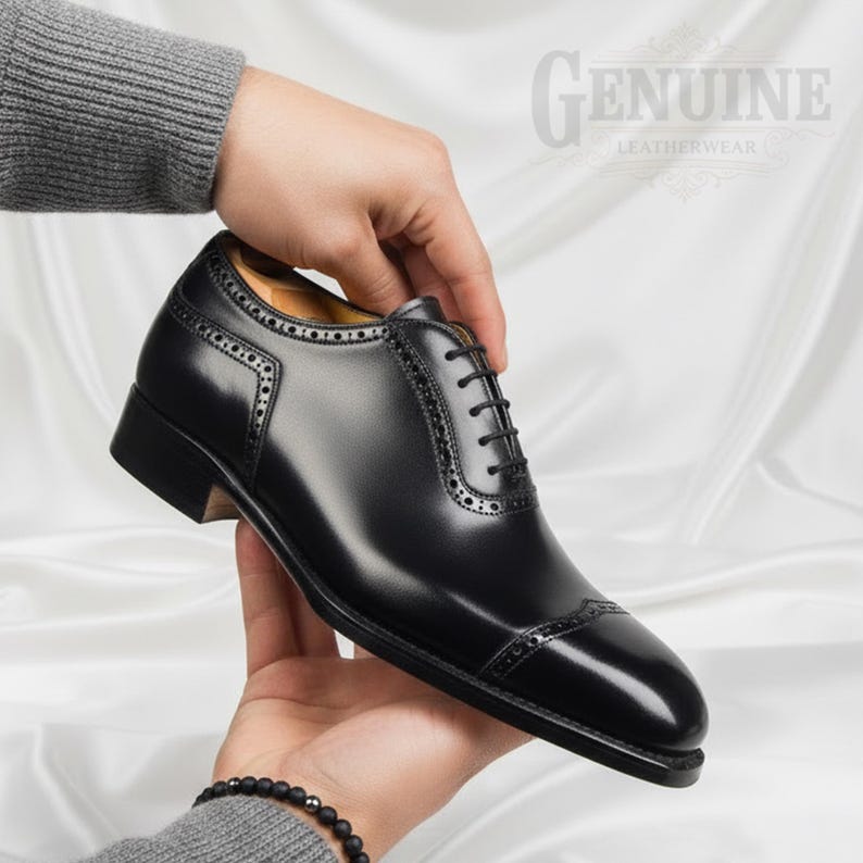 Puede incluir: Un zapato oxford de cuero negro con puntera decorada con perforaciones brogue a lo largo de las costuras y los paneles de los ojales. El zapato tiene un acabado pulido, cordones negros y un tac&oacute;n apilado. El texto GENUINE LEATHERWEAR es visible.