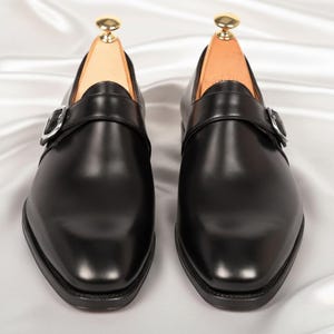 May include: A pair of black leather monk strap shoes featuring a single silver-toned metal buckle across the vamp, a rounded toe, and a low heel. Each shoe contains a light-colored wooden shoe tree with a gold-toned knob at the heel.