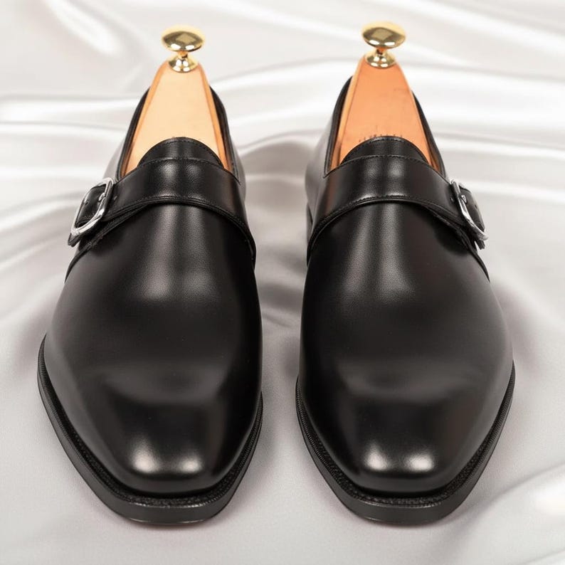 May include: A pair of black leather monk strap shoes featuring a single silver-toned metal buckle across the vamp, a rounded toe, and a low heel. Each shoe contains a light-colored wooden shoe tree with a gold-toned knob at the heel.