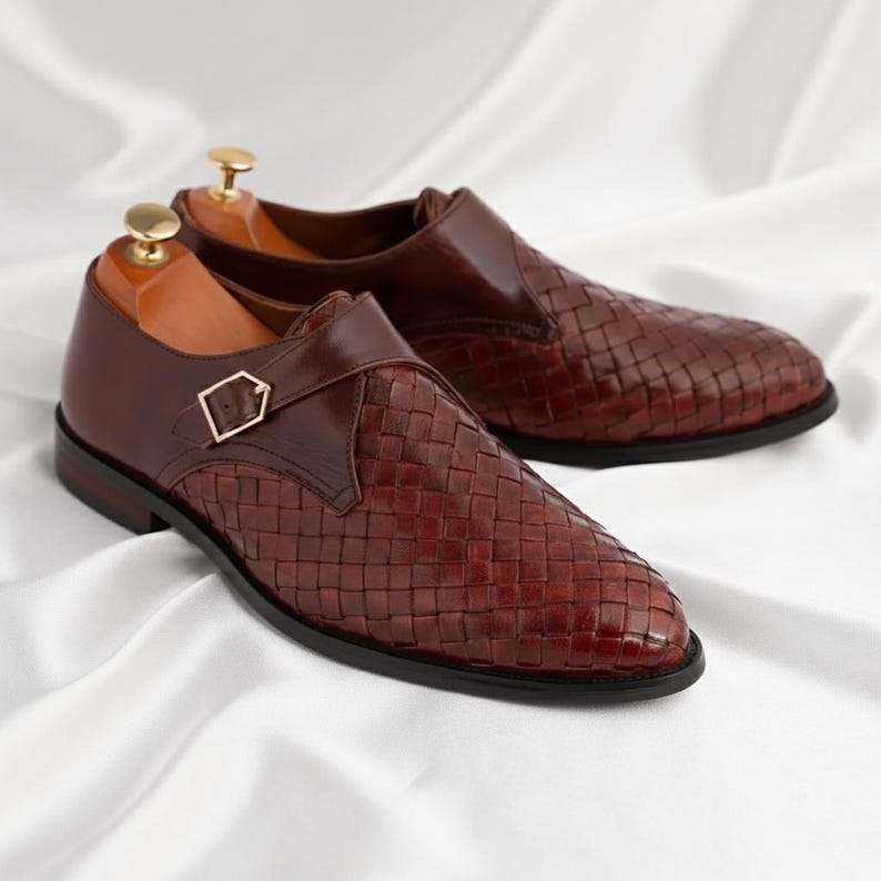 May include: A pair of dark brown leather monk strap shoes featuring a woven texture on the vamp and side panels. Each shoe has a smooth leather heel counter, a gold-toned metal buckle closure, and a wooden shoe tree inserted inside the opening.