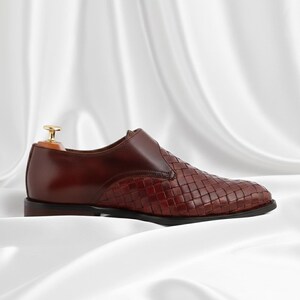 May include: A dark brown leather slip-on shoe featuring a woven texture on the front and side panels, a smooth leather heel counter, and a gold-toned metal shoe tree insert visible at the opening of the shoe, set against a white fabric background.