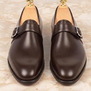 May include: A pair of dark brown leather monk strap shoes featuring a single silver-toned metal buckle closure on the side of each shoe. The shoes have a smooth finish, a rounded toe, and are equipped with light-colored wooden shoe trees inside.