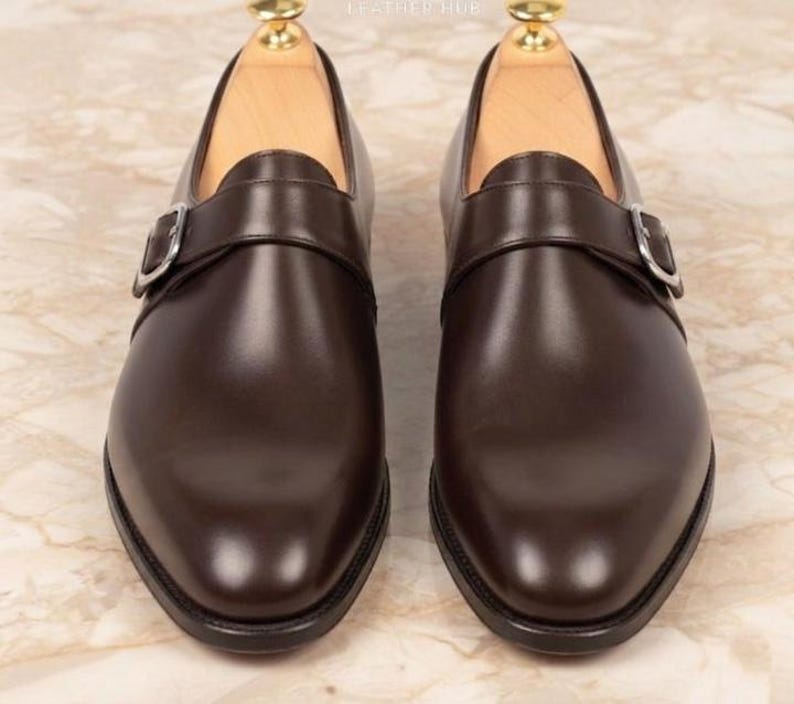 May include: A pair of dark brown leather monk strap shoes featuring a single silver-toned metal buckle closure on the side of each shoe. The shoes have a smooth finish, a rounded toe, and are equipped with light-colored wooden shoe trees inside.