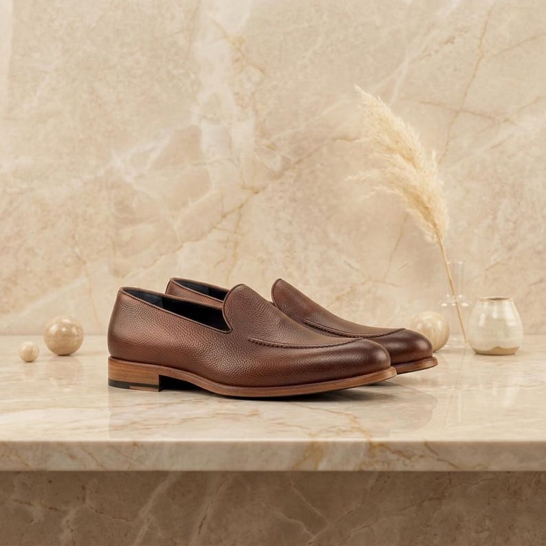 May include: A pair of brown leather loafers with a textured finish. The shoes have a classic design with a low heel and a rounded toe. The shoes are displayed on a marble surface.