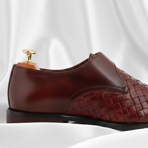 May include: A dark brown leather shoe featuring a woven texture on the front section and a smooth leather finish on the heel. The shoe includes a gold-toned metal shoe tree insert at the back, a low heel, and a dark sole, presented in a side profile.